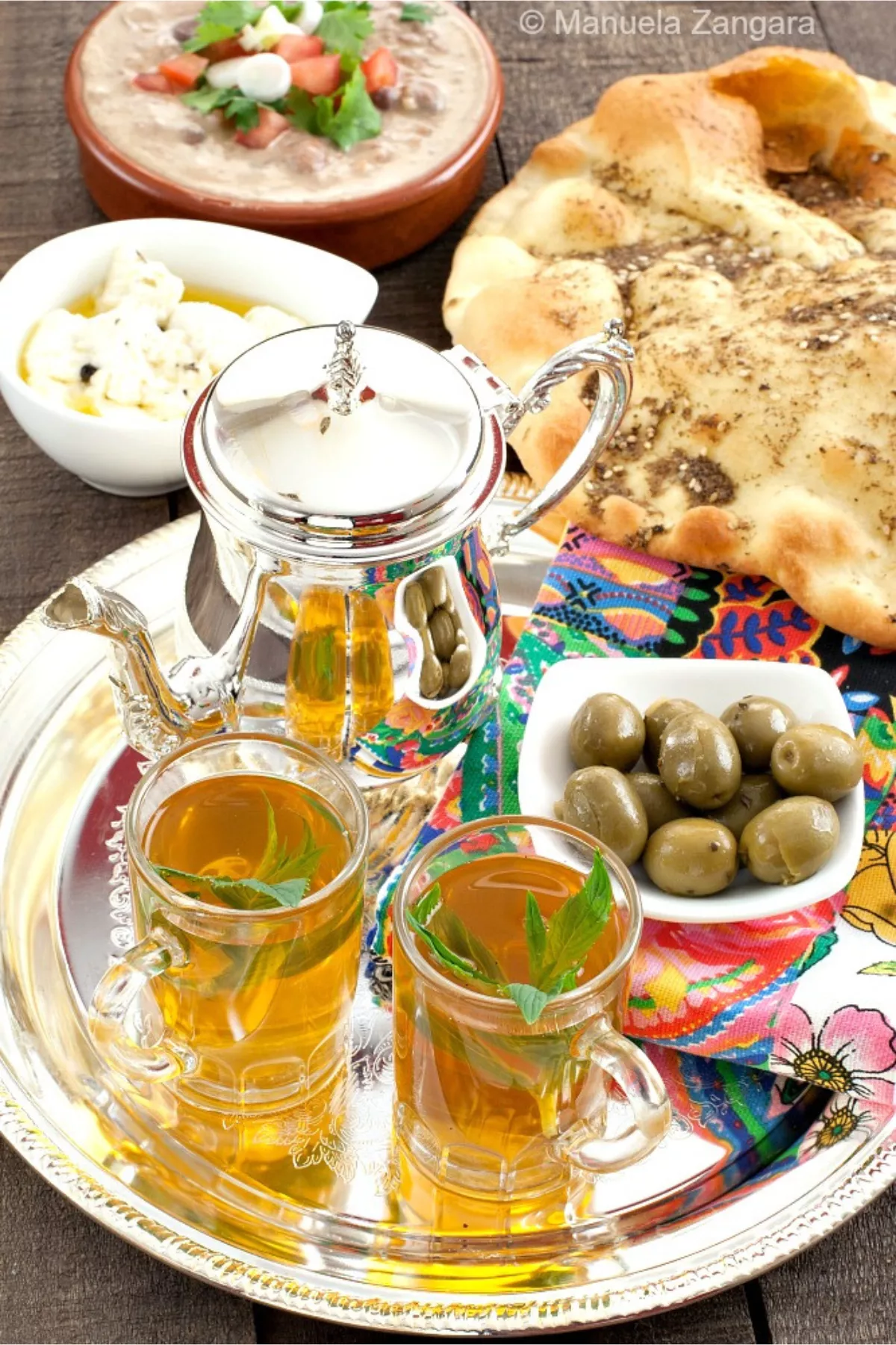 Moroccan mint tea served in glasses with mint leaves on a silver tray beside flatbread, olives, and dips.