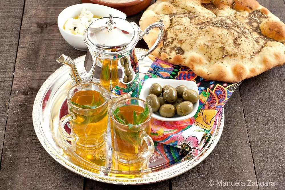 Two glasses of Moroccan mint tea with mint leaves on a tray beside olives and flatbread.