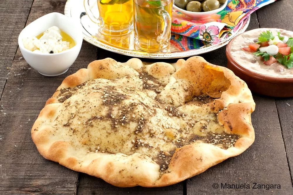 Za’atar Manouche Lebanese flatbread served with mint tea, olives, and cheese on a wooden table.