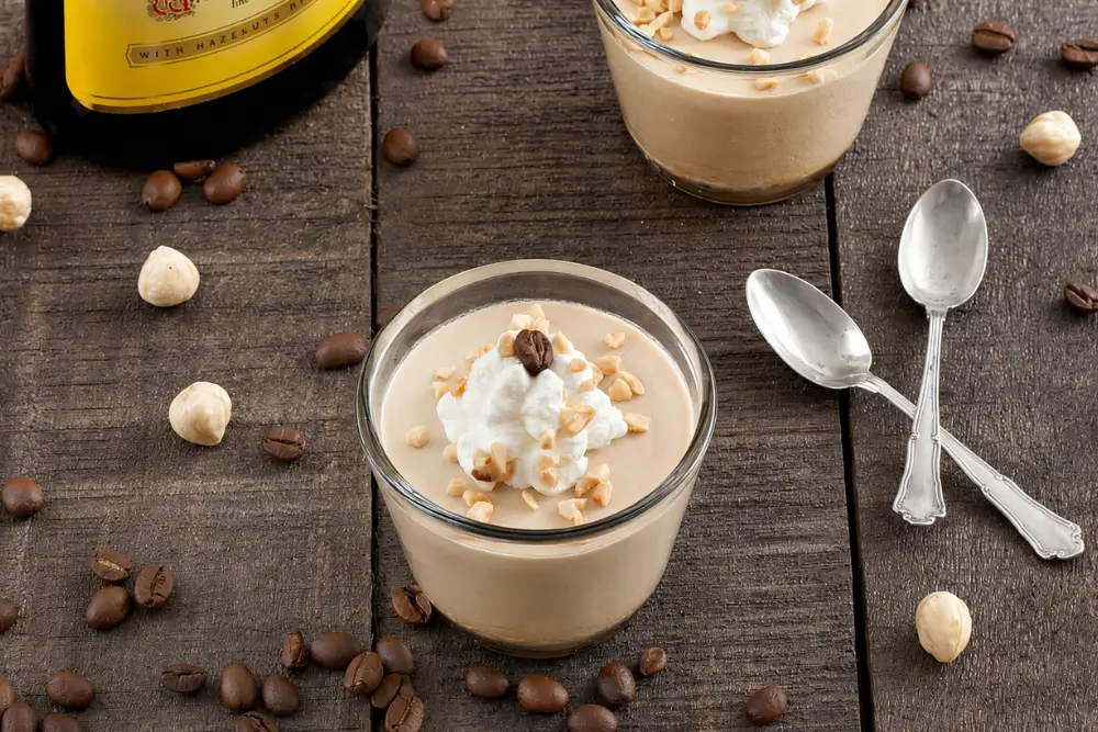 Chilled coffee mousse served in glasses with whipped cream, chopped hazelnuts, coffee beans, and spoons.