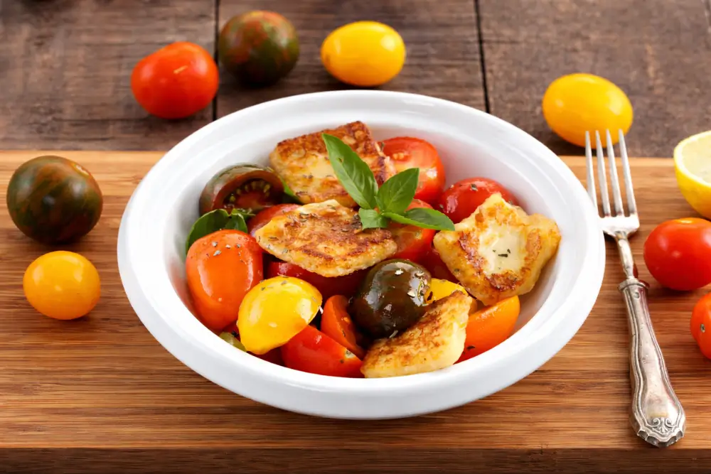 Bowl of halloumi and tomato salad served on a wooden board with fresh ingredients around.