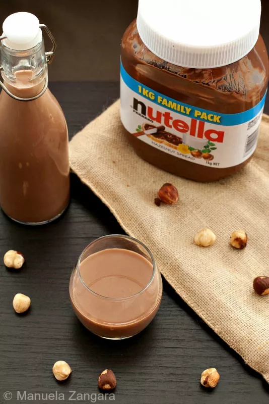 Glass and bottle of Nutella liqueur with hazelnuts on dark table.