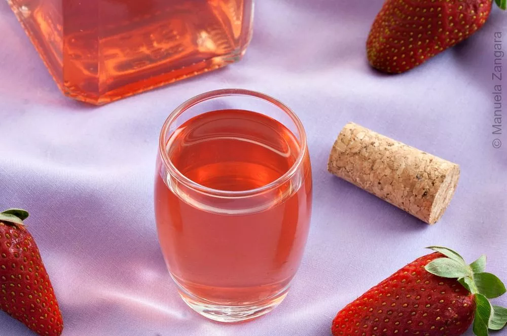 Fragolino Liqueur in a clear glass with fresh strawberries and cork on a lilac cloth.