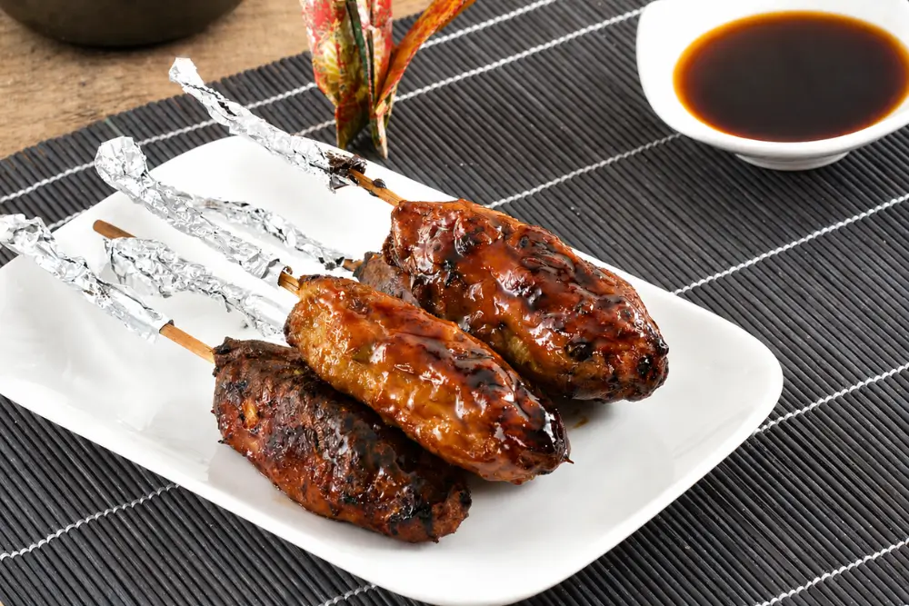Yakitori-glazed chicken skewers with charred edges served with dipping sauce on a white platter.
