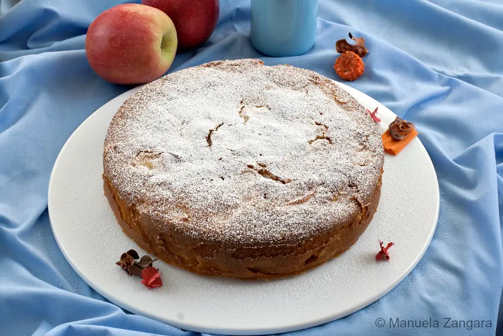 A whole apple cake dusted with icing sugar before slicing.