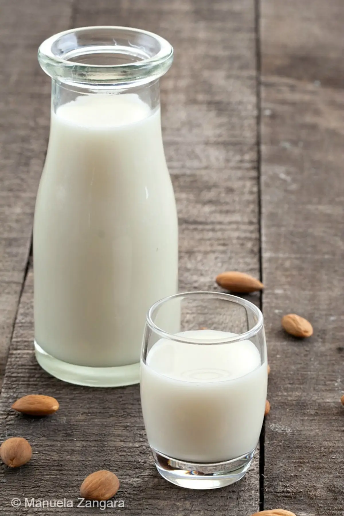 Small glass of almond milk with whole almonds scattered on a wooden surface.
