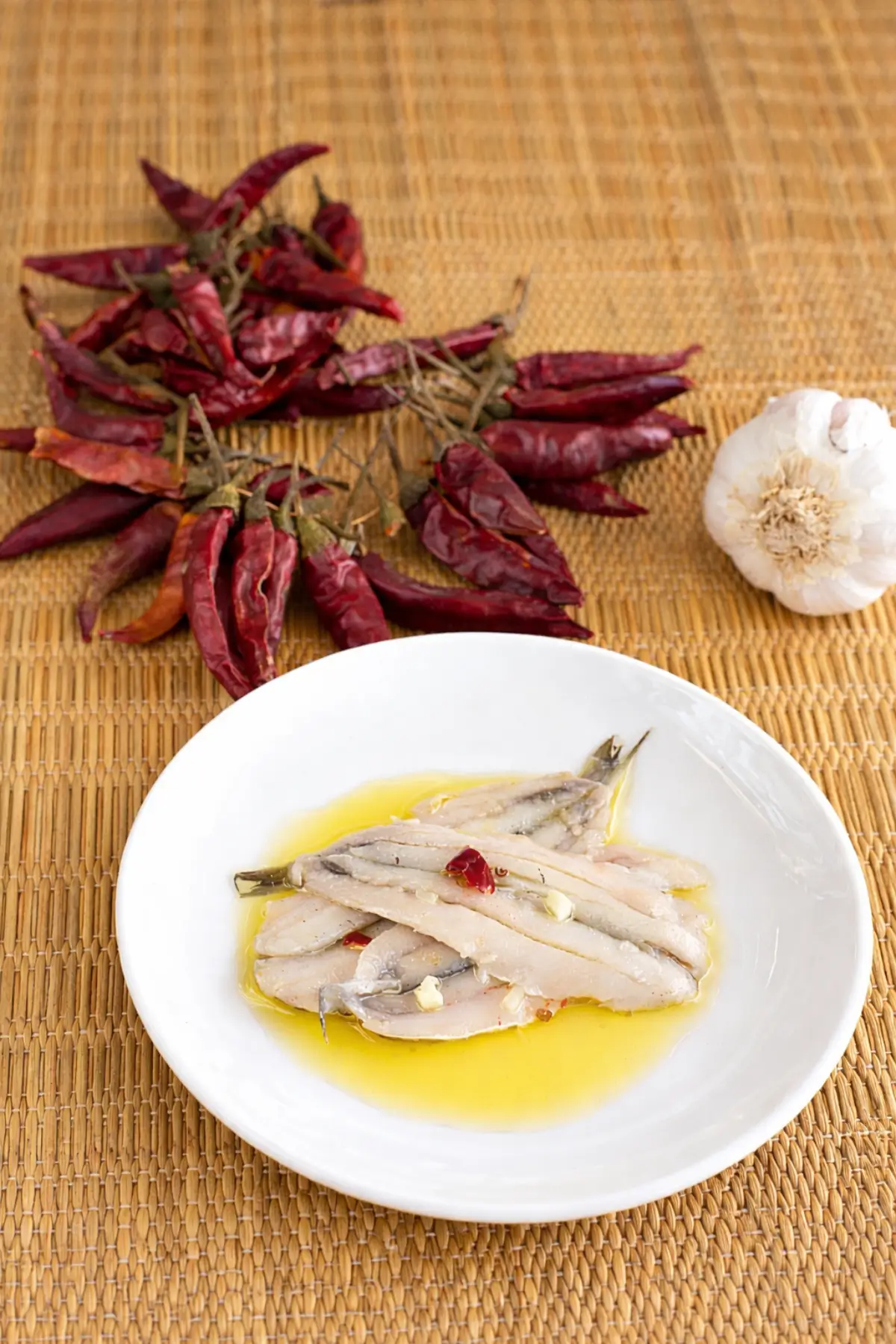 Anchovy fillets plated with olive oil, garlic pieces, and dried chilli on a white dish.
