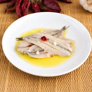 Marinated anchovies served with olive oil, garlic pieces, and chilli flakes in a shallow white bowl.
