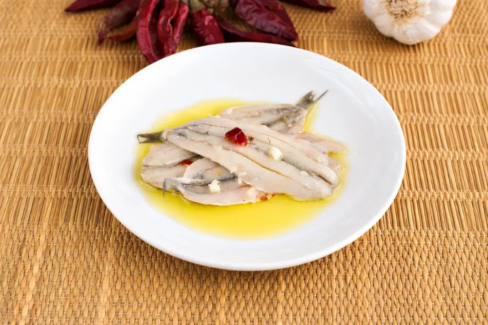 Marinated anchovies served with olive oil, garlic pieces, and chilli flakes in a shallow white bowl.