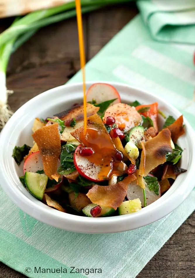 Fattoush salad with toasted pita, fresh vegetables, and sumac dressing being poured over.