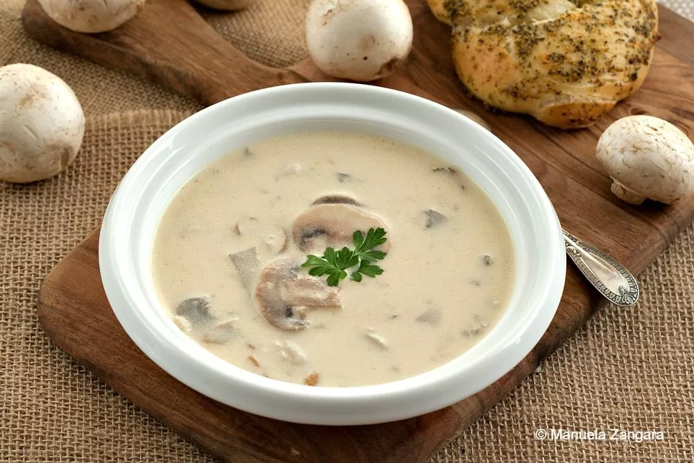 Bowl of homemade cream of mushroom soup with sliced mushrooms and a parsley garnish, served beside garlic bread.