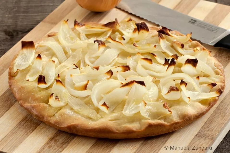 Whole Onion Schiacciata on a wooden board with onions and knife beside it.