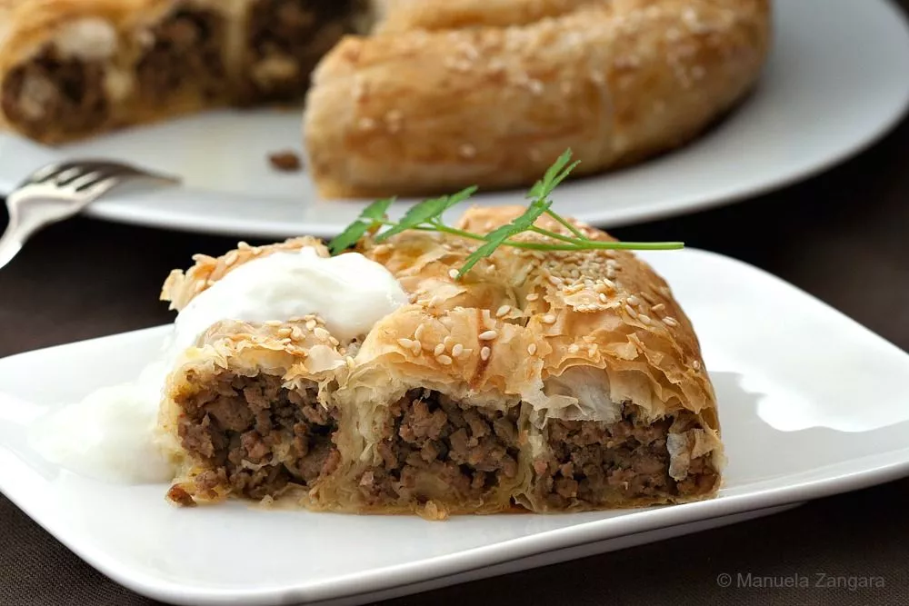 Golden brown baked Bosnian burek spiral topped with sesame seeds and served with yoghurt.