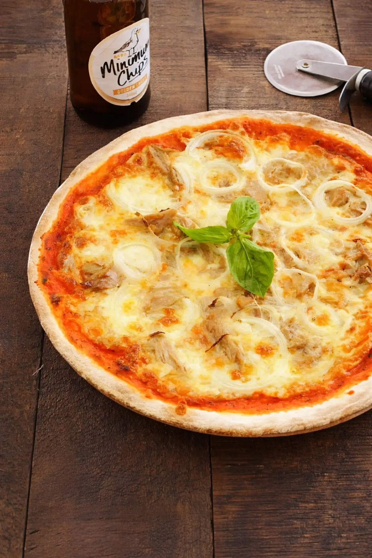 Tuna and onion pizza served on a rustic wooden table with a pizza cutter and bottle in the background.