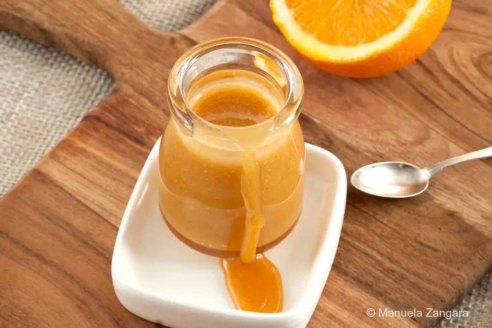 Top view of a jar filled with caramel sauce with orange on a wooden board with a spoon and halved orange beside it.