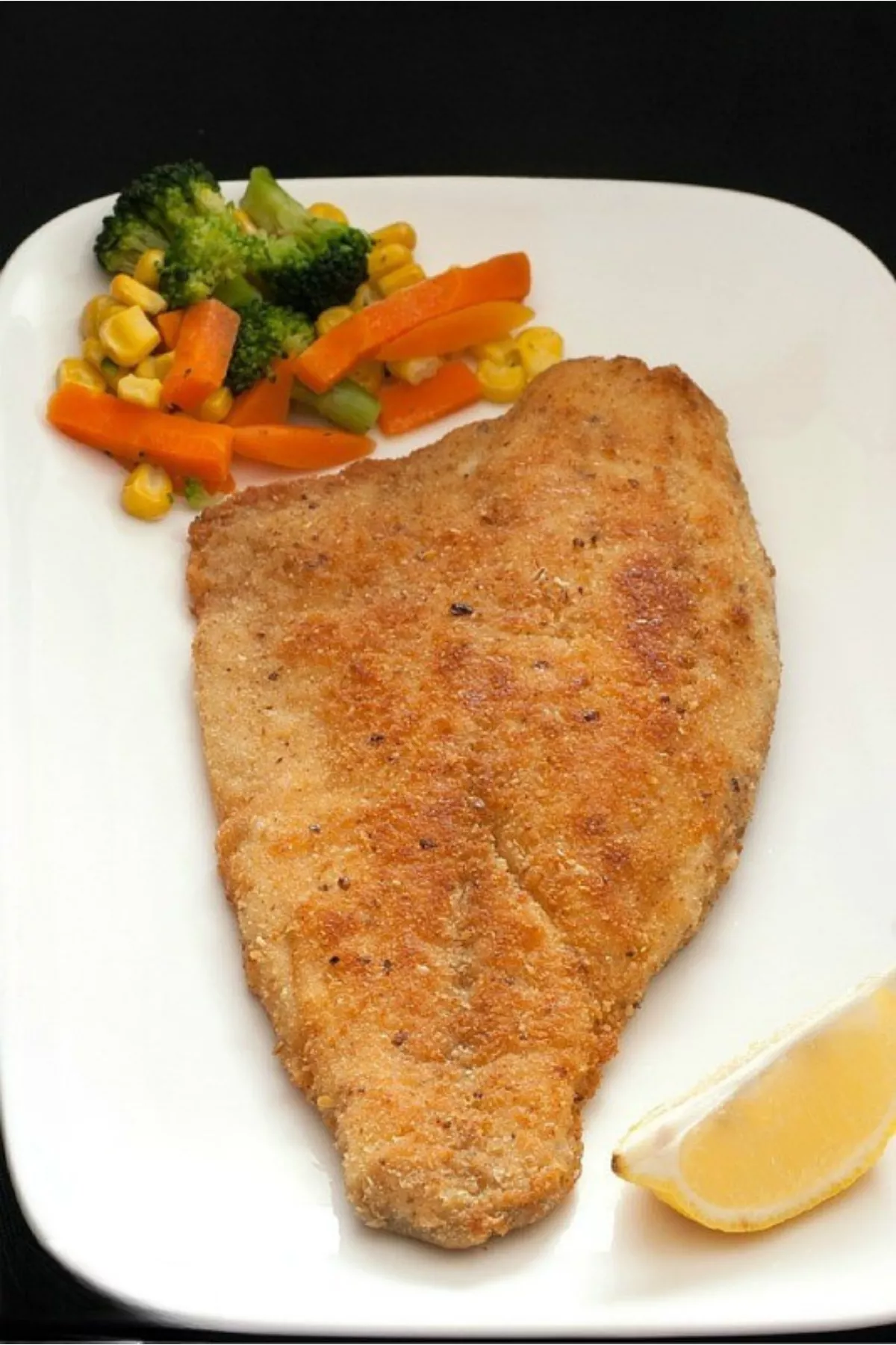 A plate of Crumbed Barramundi served with mixed vegetables and a wedge of lemon.