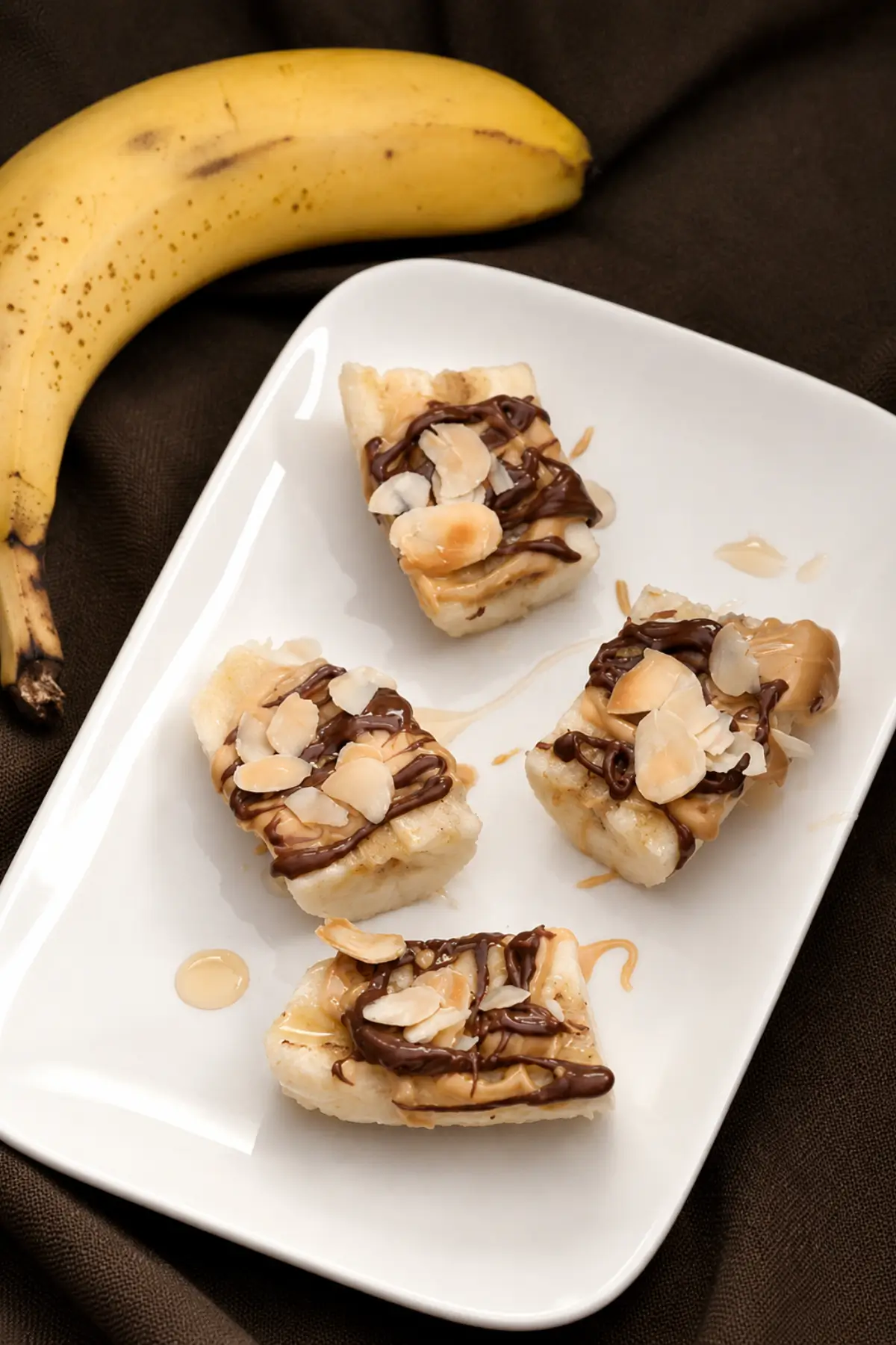 Banana bites with Nutella, peanut butter, honey, and almond slices served on a white plate.