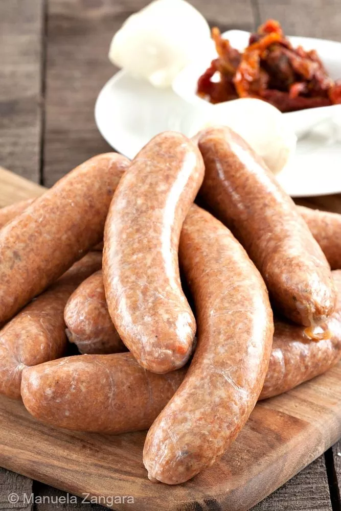 Mozzarella and Sun-Dried Tomato Pork Sausages on a wooden board with cheese and tomatoes in the background.