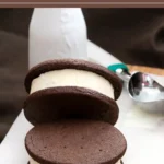 Chocolate cookie ice cream sandwich with thick vanilla filling served on parchment paper with an ice cream scoop beside it.