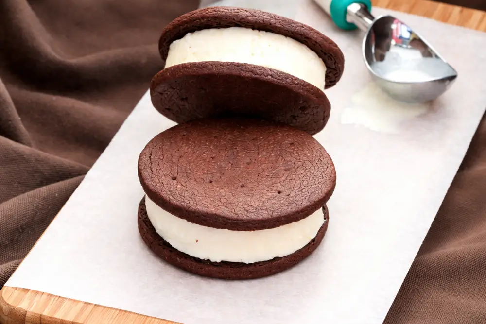 Chocolate cookie ice cream sandwiches stacked with thick vanilla filling on parchment paper next to an ice cream scoop.