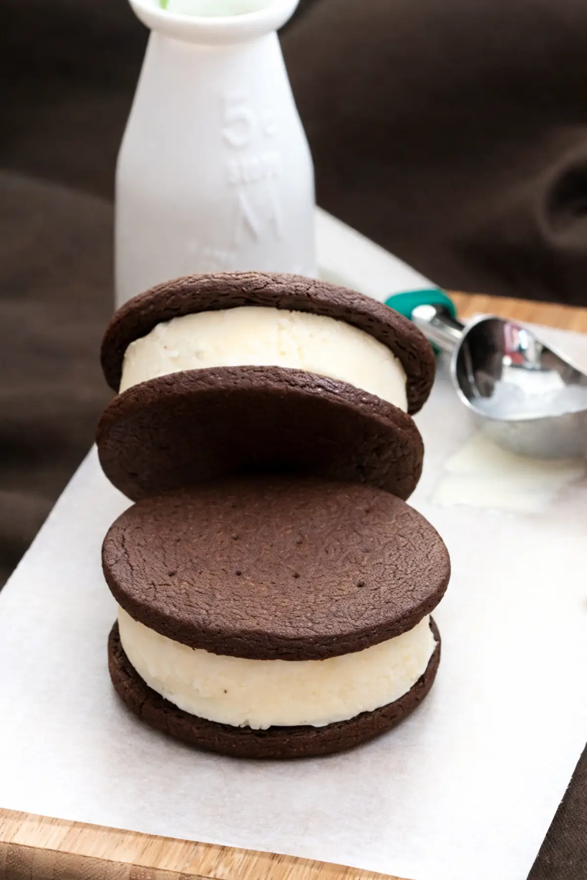 Chocolate cookie ice cream sandwich with thick vanilla filling served on parchment paper with an ice cream scoop beside it.