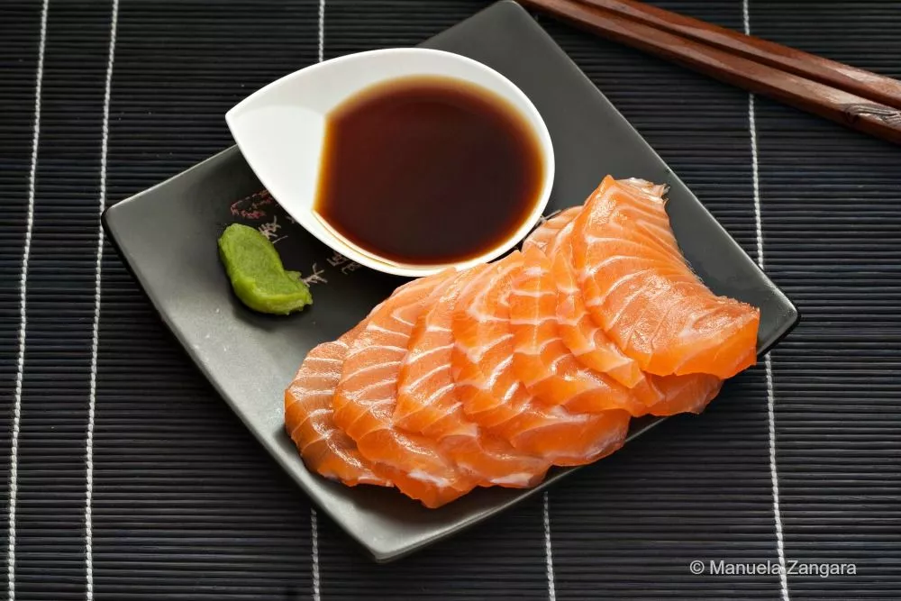 Close-up of sliced salmon sashimi on a black plate with Ponzu sauce and wasabi.