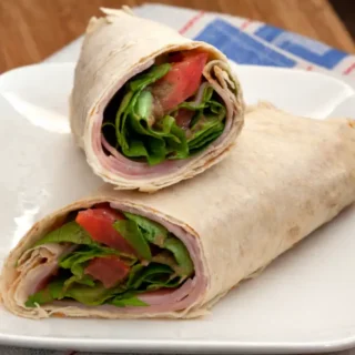 Two halves of a ham salad wrap with fresh lettuce and tomato served on a white plate.