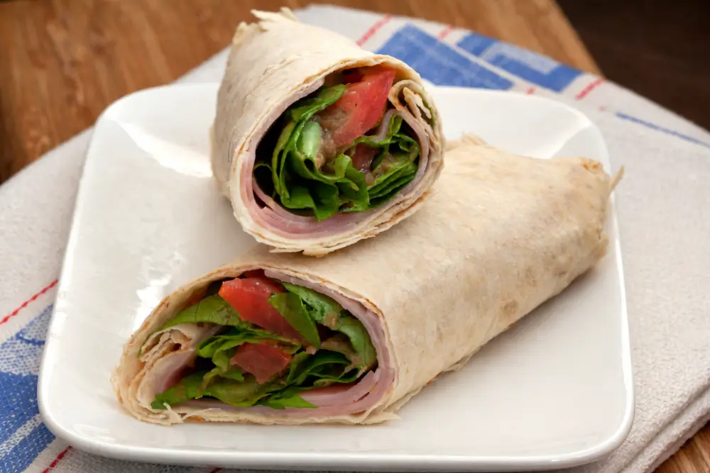 Two halves of a ham salad wrap with fresh lettuce and tomato served on a white plate.