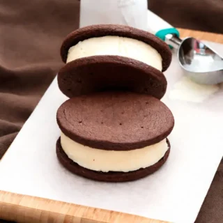 Chocolate cookie ice cream sandwiches stacked with vanilla filling on parchment paper.