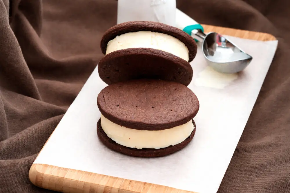 Chocolate cookie ice cream sandwiches stacked with vanilla filling on parchment paper.