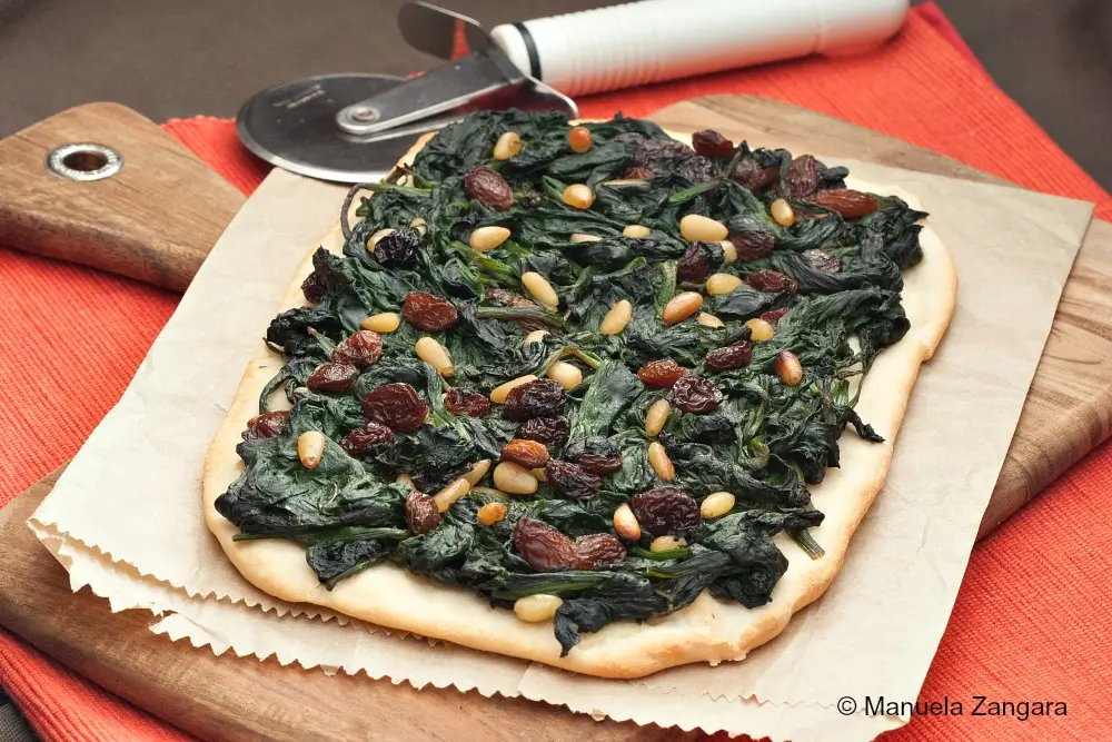 Baked Coca de Espinacas a la Catalana topped with spinach, raisins, and pine nuts on a wooden board.