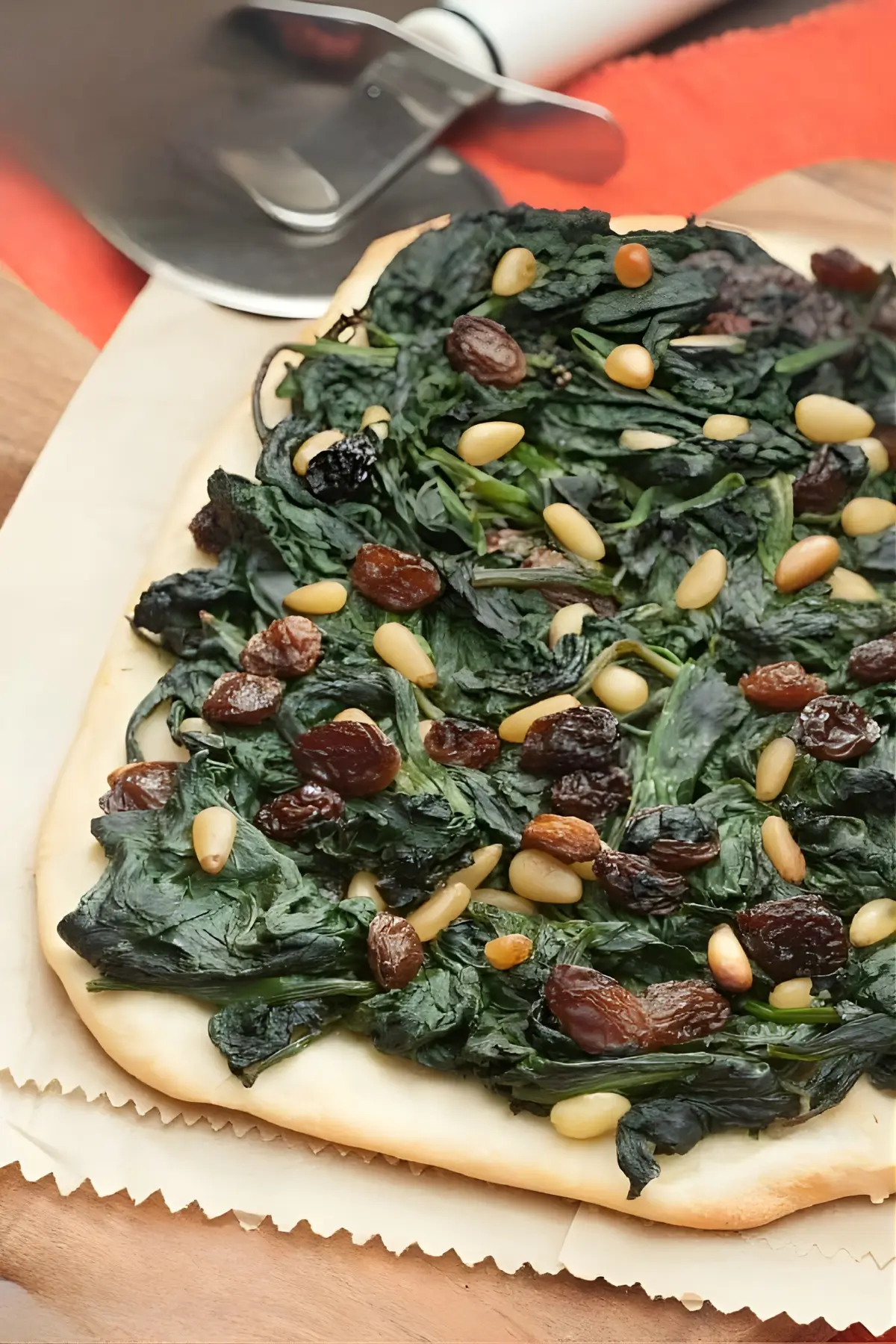 Coca de Espinacas a la Catalana topped with spinach, raisins, and pine nuts on baking paper over a wooden board.