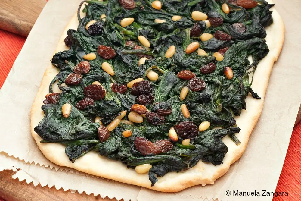 Close-up of baked coca topped with spinach, raisins, and pine nuts.