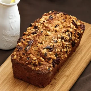 Freshly baked banana bread topped with mixed nuts beside a milk bottle.