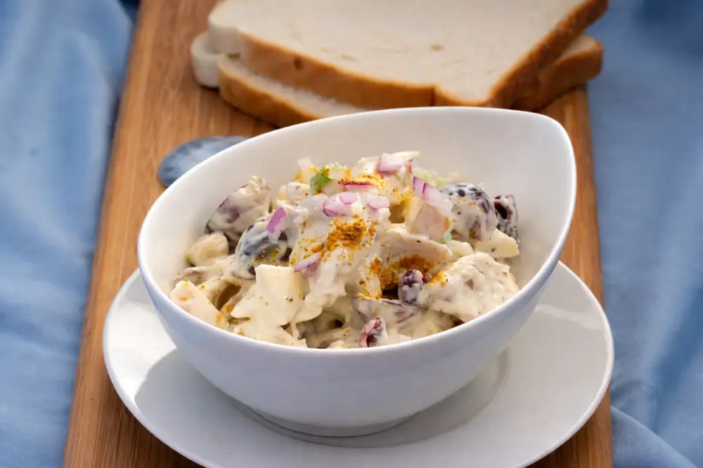 Creamy chicken salad in a bowl placed on a wooden board with sliced sandwich bread in the background.