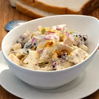 Curried chicken salad served in a white bowl with grapes, apples, and a creamy dressing, topped with a sprinkle of curry powder.