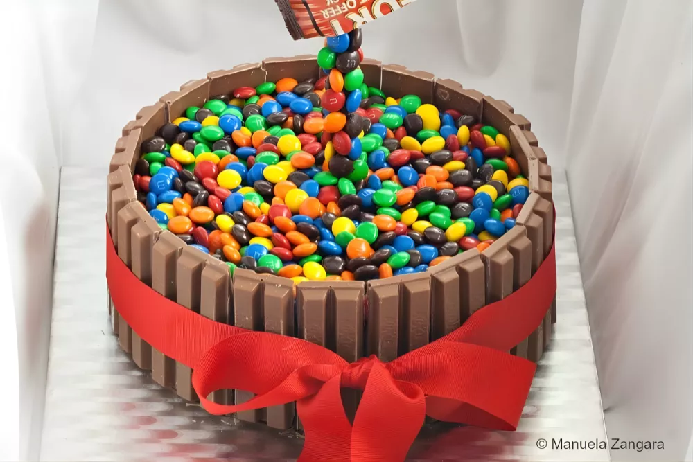 Finished M&M’s Kit Kat cake with a red ribbon and a floating candy effect.