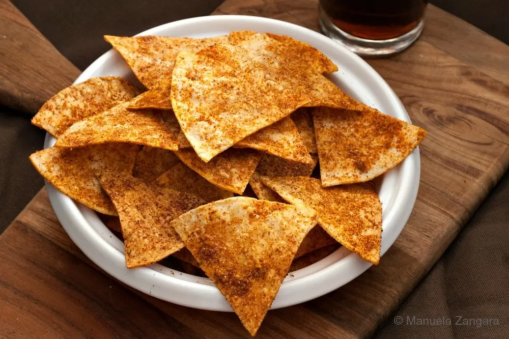Homemade Doritos in a white bowl on a wooden board with a drink in the background.