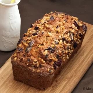 Whole banana nut bread topped with chopped walnuts and pecans beside a milk jar on a wooden board.