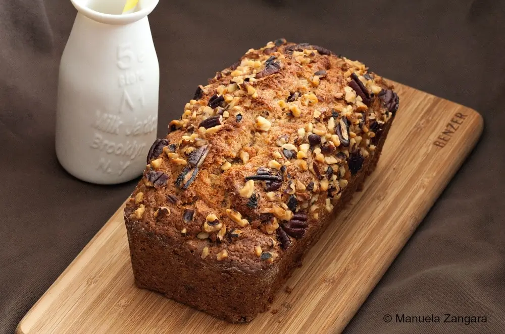 Whole banana nut bread topped with chopped walnuts and pecans beside a milk jar on a wooden board.