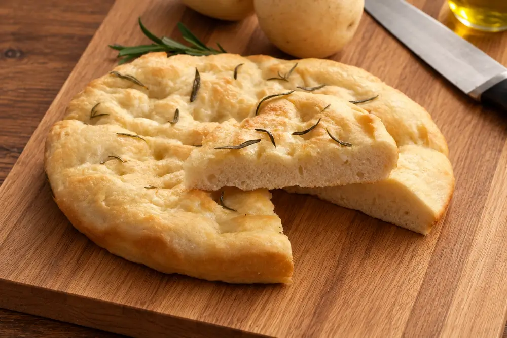Sliced rosemary focaccia with a soft crumb beside whole potatoes.