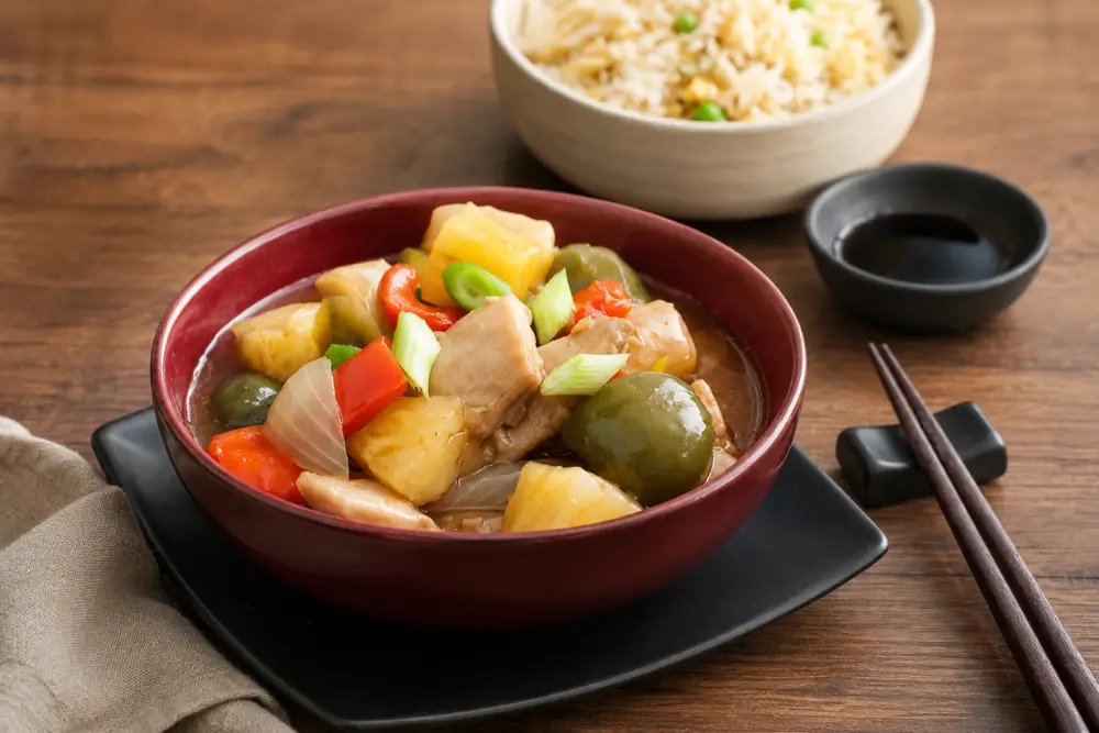 Pineapple chicken stir-fry served in a red bowl with fried rice and soy sauce.