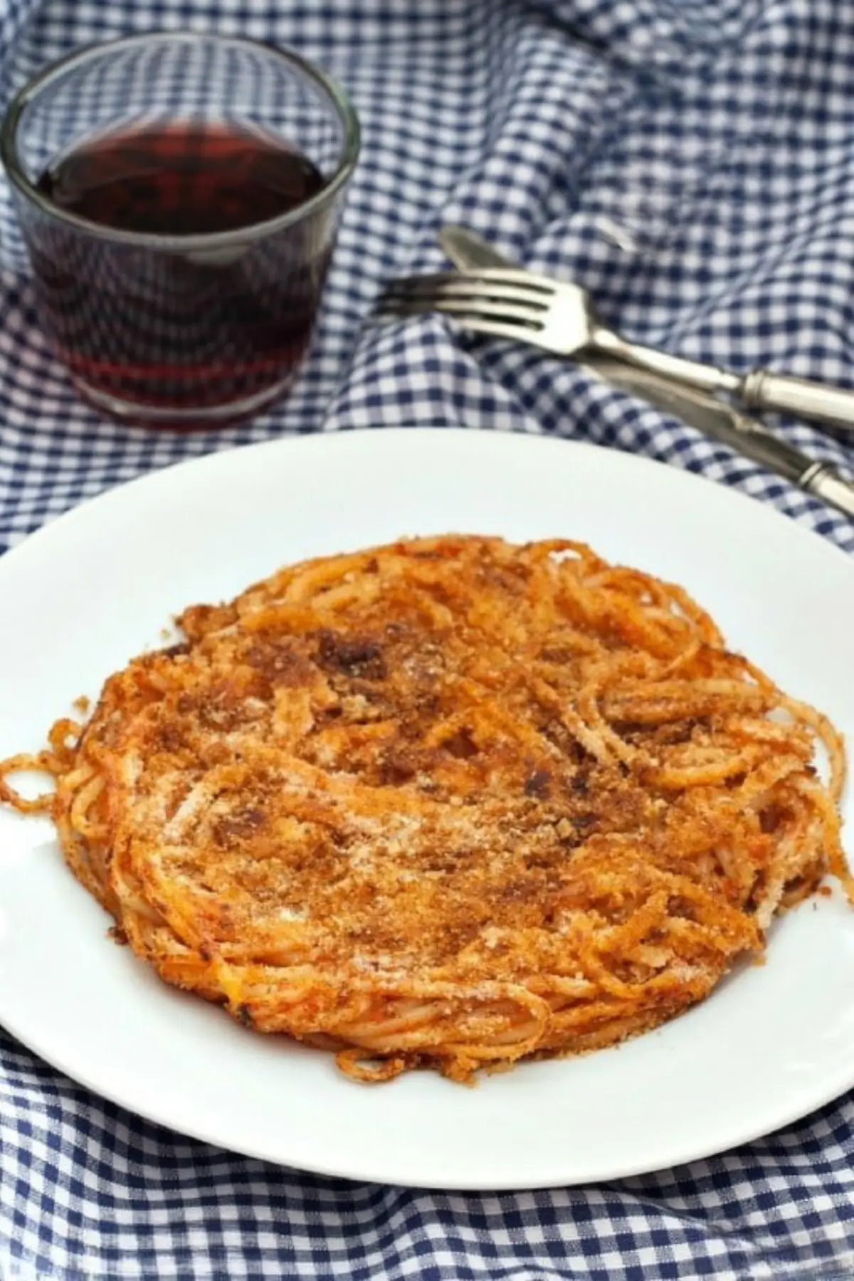 Golden fried spaghetti cake served on a white plate with a glass of red wine and fork on a blue checkered cloth.