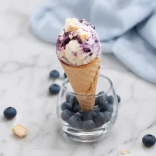 Scoop of Blueberry Cheesecake Ice Cream in a waffle cone set in a glass filled with fresh blueberries.