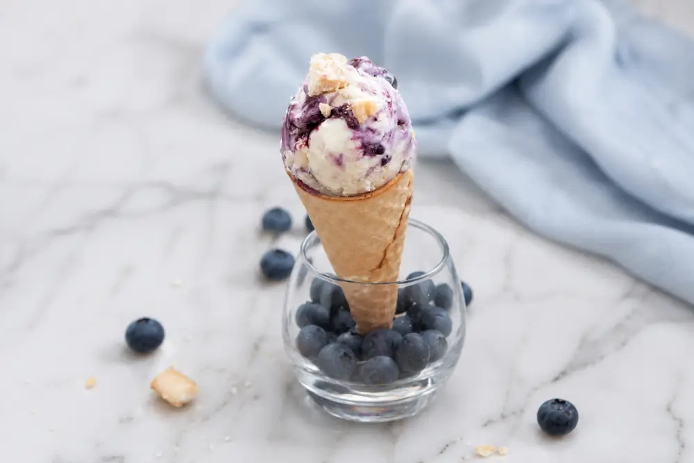 Scoop of Blueberry Cheesecake Ice Cream in a waffle cone set in a glass filled with fresh blueberries.