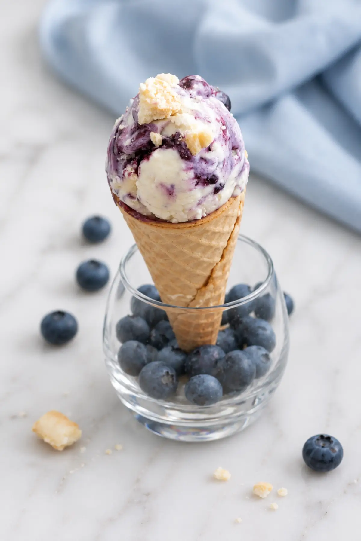 Homemade blueberry cheesecake ice cream served in a waffle cone with fresh blueberries and cookie crumbs.