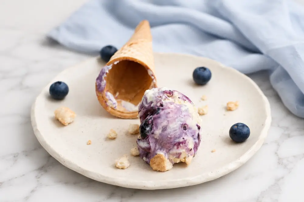 Scoop of blueberry cheesecake ice cream with cookie crumbs beside a fallen waffle cone on a plate.
