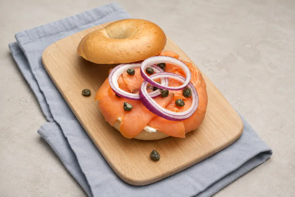 Open bagel topped with salmon, red onion and capers on a wooden board.