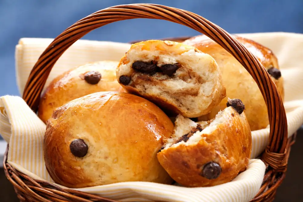 Golden baked chocolate chip buns in a basket, one cut open with melted chocolate.