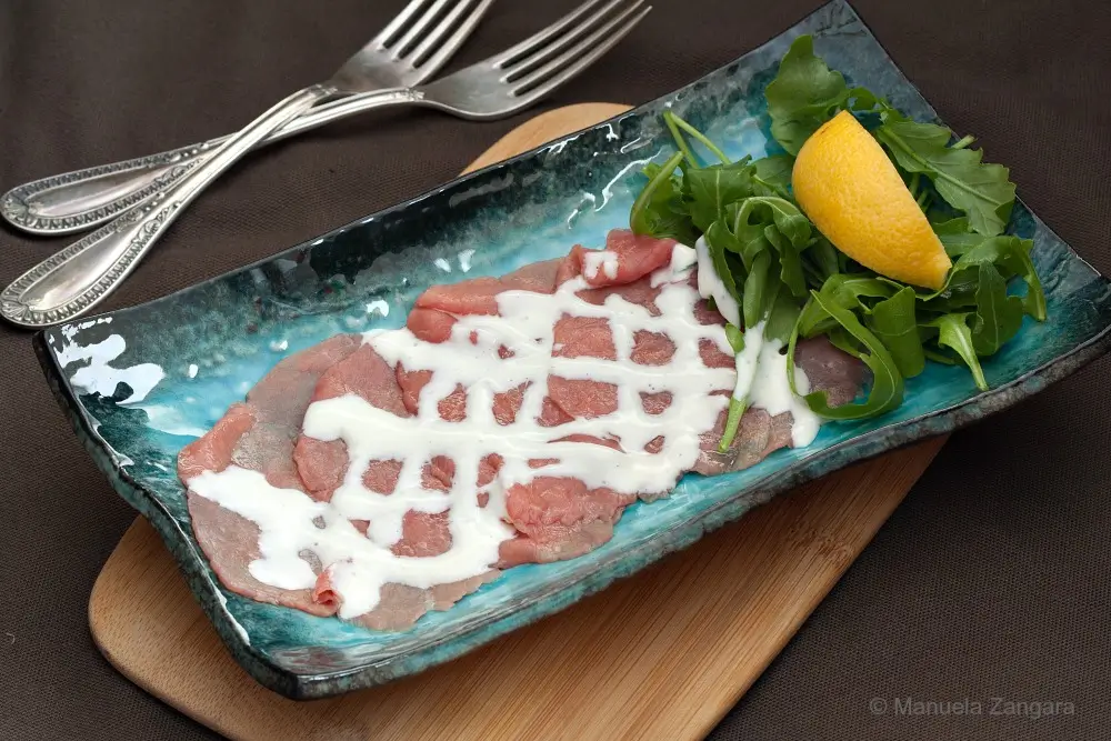 Close-up view of carpaccio drizzled with Cipriani sauce beside rocket and a lemon wedge.